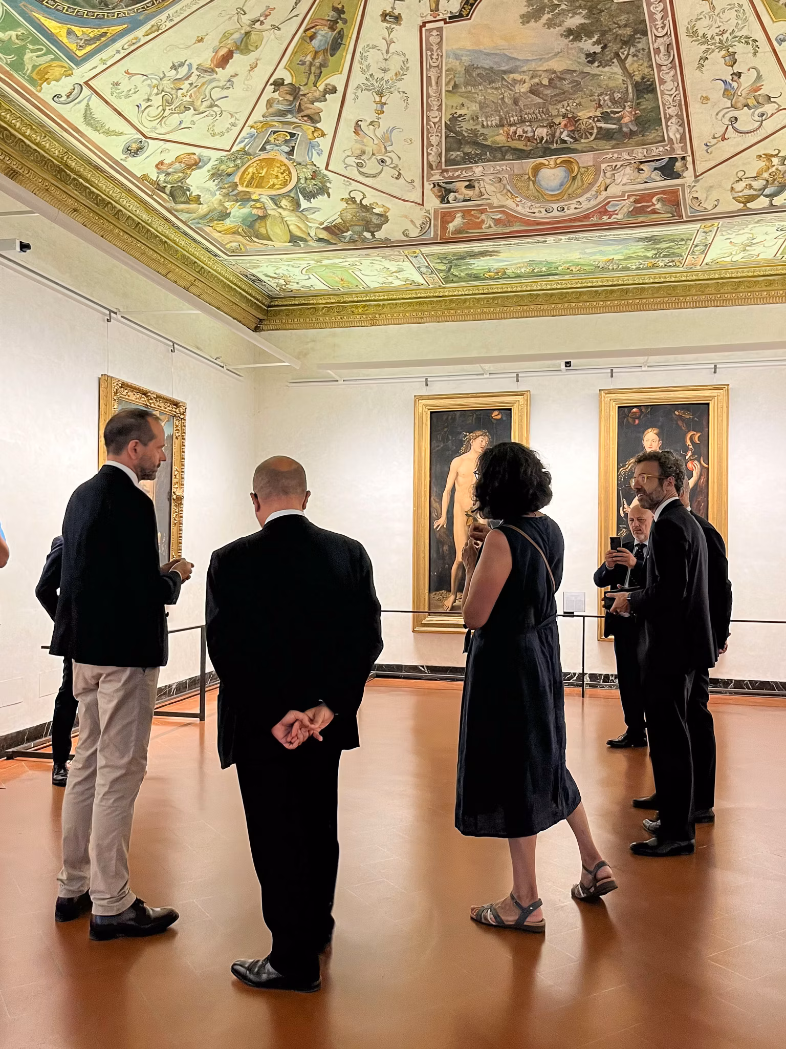 Il Ministro della Cultura Gennaro Sangiuliano in visita agli Uffizi nelle sale dei maestri fiamminghi Un gruppo di persone osserva attentamente dipinti rinascimentali, incorniciati su pareti bianche, sotto un soffitto decorato riccamente con affreschi storici all'interno di una galleria d'arte.