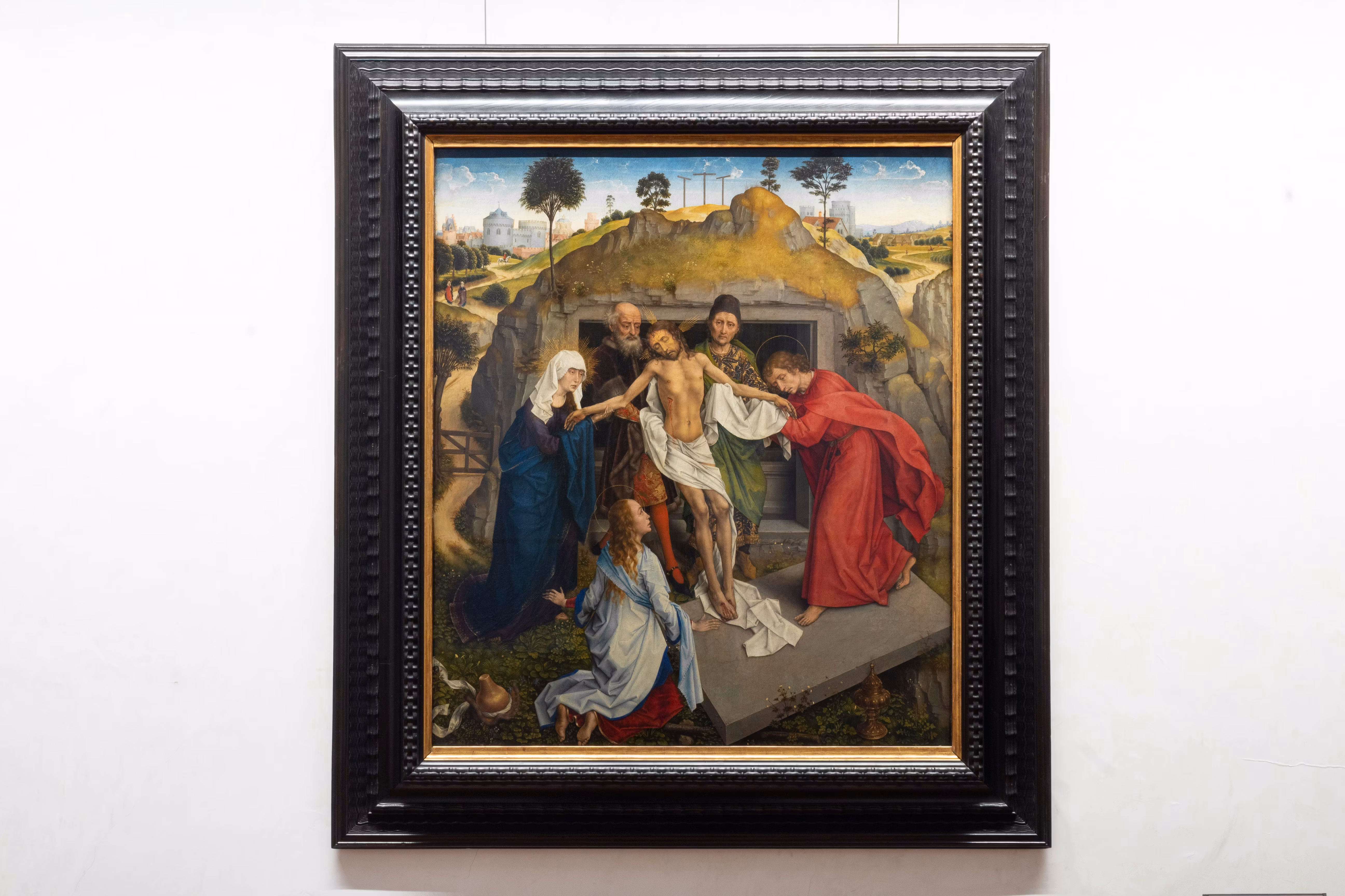 Un dipinto incorniciato raffigura la deposizione di Cristo dalla croce, circondato da figure vestite in abiti tradizionali, ambientato in un paesaggio rurale con alberi e colline sullo sfondo.